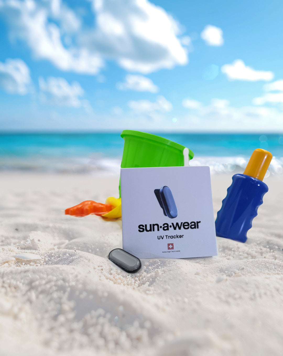 Sandy beach on a hot day with sun-a-wear UV tracker in focus in front of other beach equipment